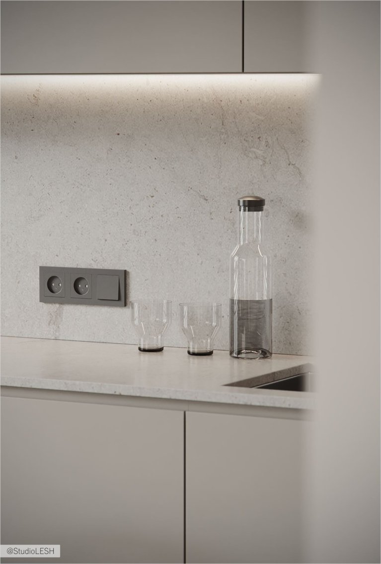 На фотографии изображена часть кухонного интерьера с серыми шкафами, светлой столешницей, двумя стаканами, бутылкой с водой и электрическими розетками. Виден также край раковины