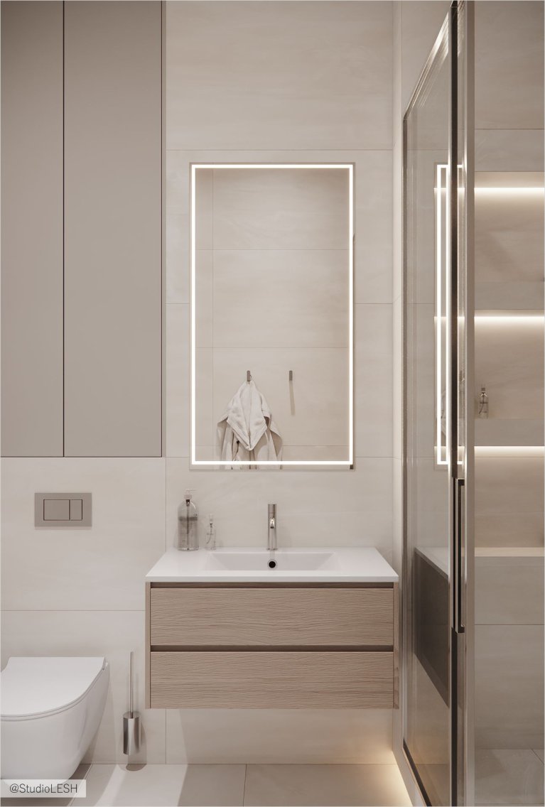 Интерьер ванной комнаты в светлых тонах с душевой кабиной, частично огороженной стеклом, подвесной тумбой с раковиной, зеркалом с подсветкой и подвесным унитазом. На стене висит белое полотенце