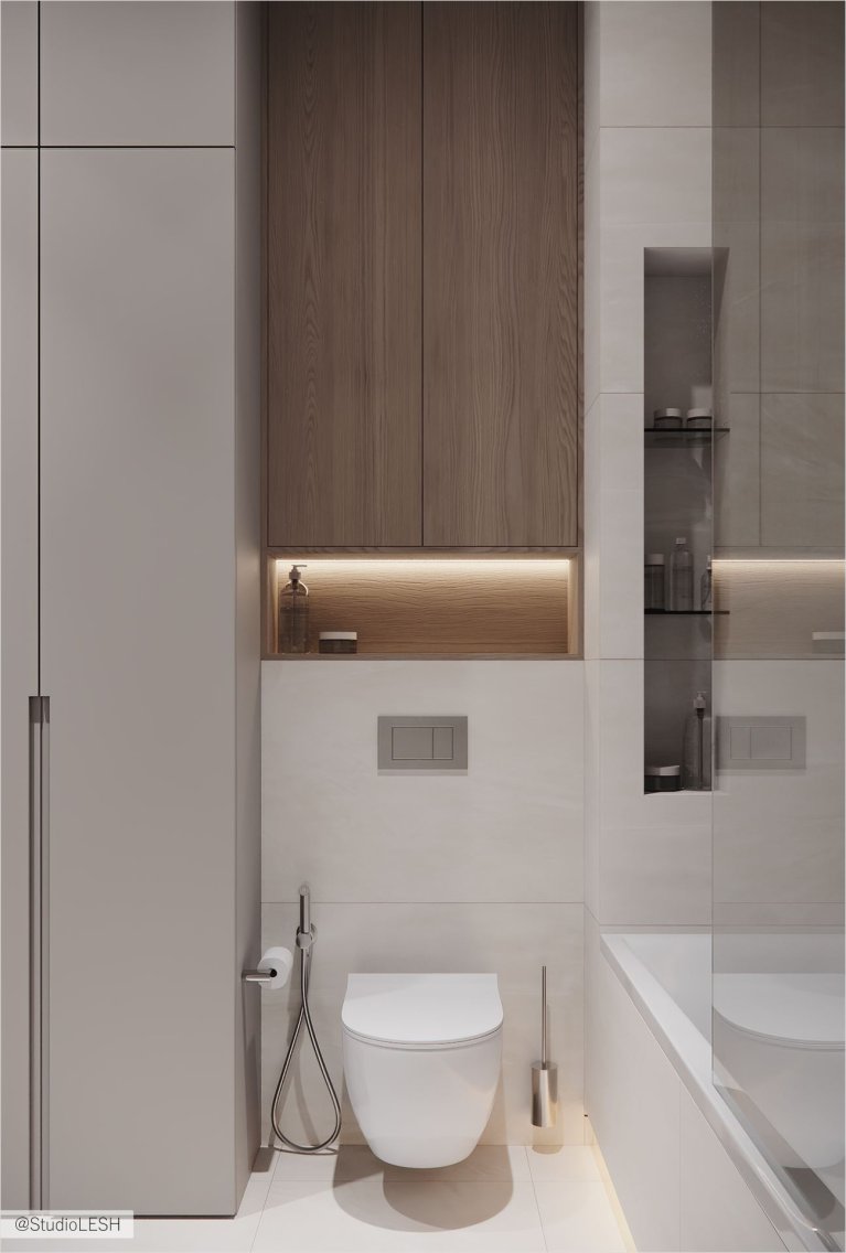 Интерьер ванной комнаты в светлых тонах с подвесным унитазом, гигиеническим душем, душевой кабиной, частично огороженной стеклом, и встроенным шкафчиком с деревянными полками. На заднем плане - серые стены и белая сантехника