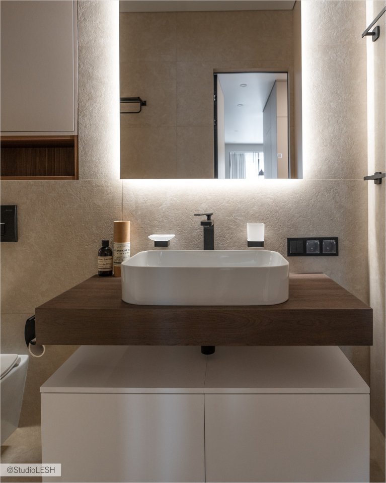 Стильная ванная комната с подвесной раковиной, установленной на столешнице из темного дерева. Над раковиной висит большое зеркало с подсветкой, отражающее светлые стены и полки с аксессуарами. Видны также черные смесители и электрические розетки