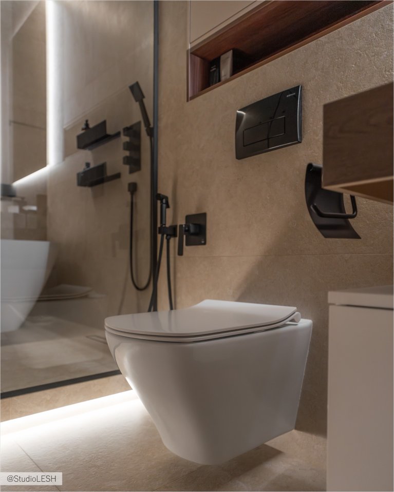 Интерьер ванной комнаты в светлых тонах с подвесным унитазом, гигиеническим душем, душевой кабиной, частично огороженной стеклом, и встроенным шкафчиком с деревянными полками. На заднем плане - серые стены и белая сантехника