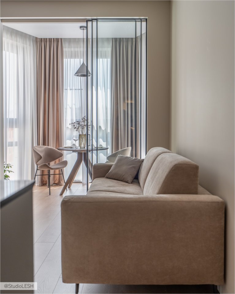 Интерьер гостиной в светлых тонах с мягким диваном, обеденным столом со стульями, разделяющим пространство стеклянной перегородкой, и плотными шторами на окнах.