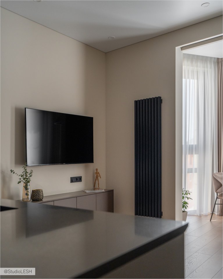 Интерьер гостиной в светлых тонах с плоским телевизором на стене, вертикальным радиатором отопления антрацитового цвета, тумбой под телевизор и минималистичным декором. Справа видна часть комнаты с окном и шторами