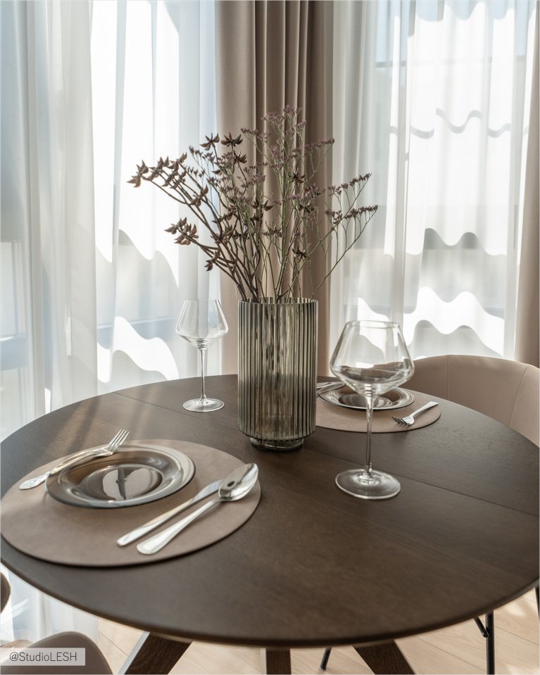 Стильный обеденный стол с темной столешницей и металлическими ножками. На столе стоят два бокала из прозрачного стекла, две тарелки с серыми салфетками и серебряные столовые приборы. В центре стола - высокая ваза из темного стекла с декоративными ветками. Окно за столом украшено плотными шторами