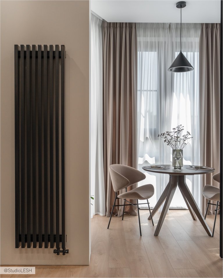Интерьер гостиной в светлых тонах с обеденным столом круглой формы, двумя стульями с мягкой обивкой, большим вертикальным радиатором отопления антрацитового цвета и плотными шторами на окнах