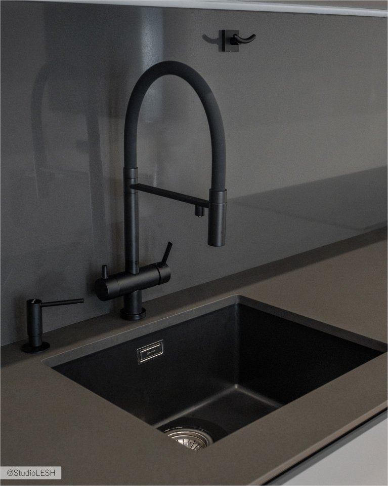 Стильная кухня с черным смесителем с гибким изливом, идеально подходящим для современной кухни. Смеситель установлен на темной столешнице рядом с темной раковиной. Видны также другие элементы кухонного гарнитура, выполненные в едином стиле, и рейлинг с крючками на заднем плане