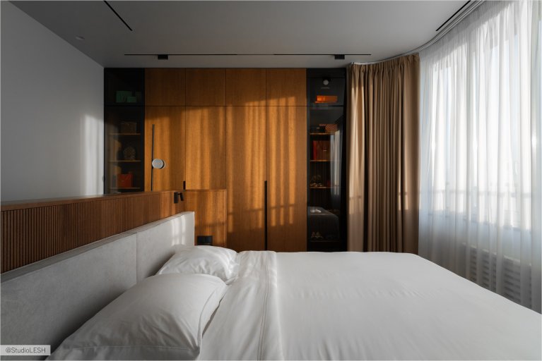 Уютная спальня с большой кроватью, застеленной белым покрывалом, и декоративными подушками. На заднем плане - вместительные деревянные шкафы с открытыми полками, на которых расставлены книги и предметы декора. Окно в комнате украшено плотными шторами, создающими атмосферу уюта и комфорта