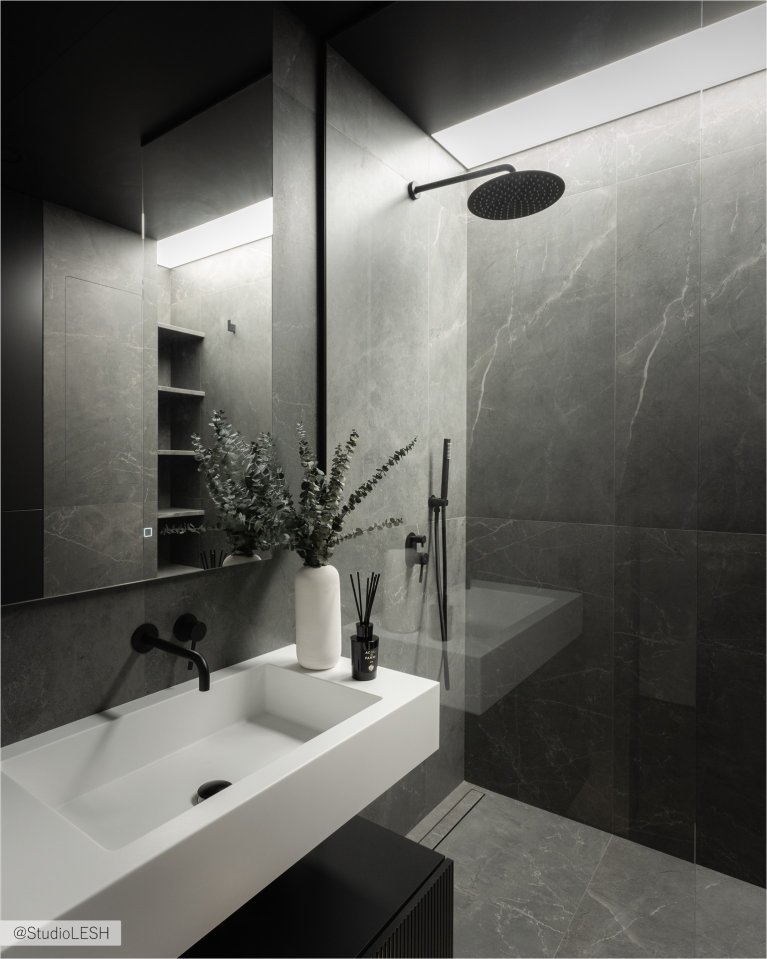 Стильная ванная комната с душевой кабиной с тропическим душем и стеклянной перегородкой. На переднем плане - подвесная тумба с раковиной и зеркалом с подсветкой. Рядом с раковиной - минималистичные аксессуары для ванной и живые растения, добавляющие уют и свежесть