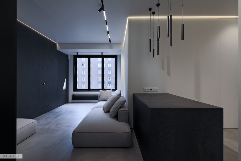 Интерьер гостиной в светлых тонах с серым диваном, большим окном с видом на город, черной стеной с подсветкой и минималистичными светильниками. В комнате также есть темный столик и другие элементы декора