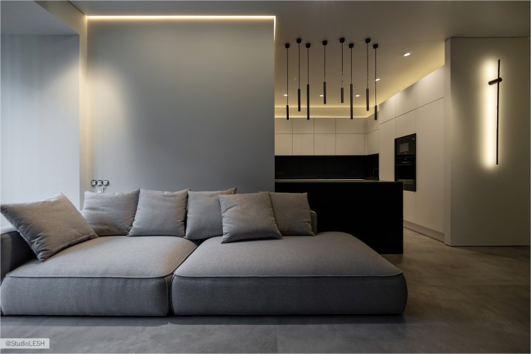 Интерьер гостиной в светлых тонах с серым диваном, большим окном с видом на город, черной стеной с подсветкой и минималистичными светильниками. В комнате также есть темный столик и другие элементы декора