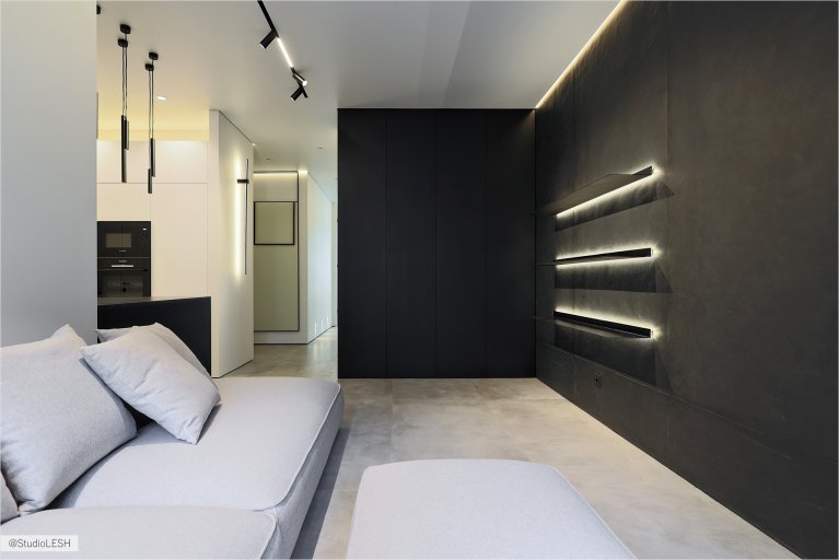 Стильная гостиная с серым диваном, украшенным подушками, и темным столиком. На заднем плане - большое окно с видом на город, черная стена с подсветкой и минималистичные светильники, создающие уютную атмосферу