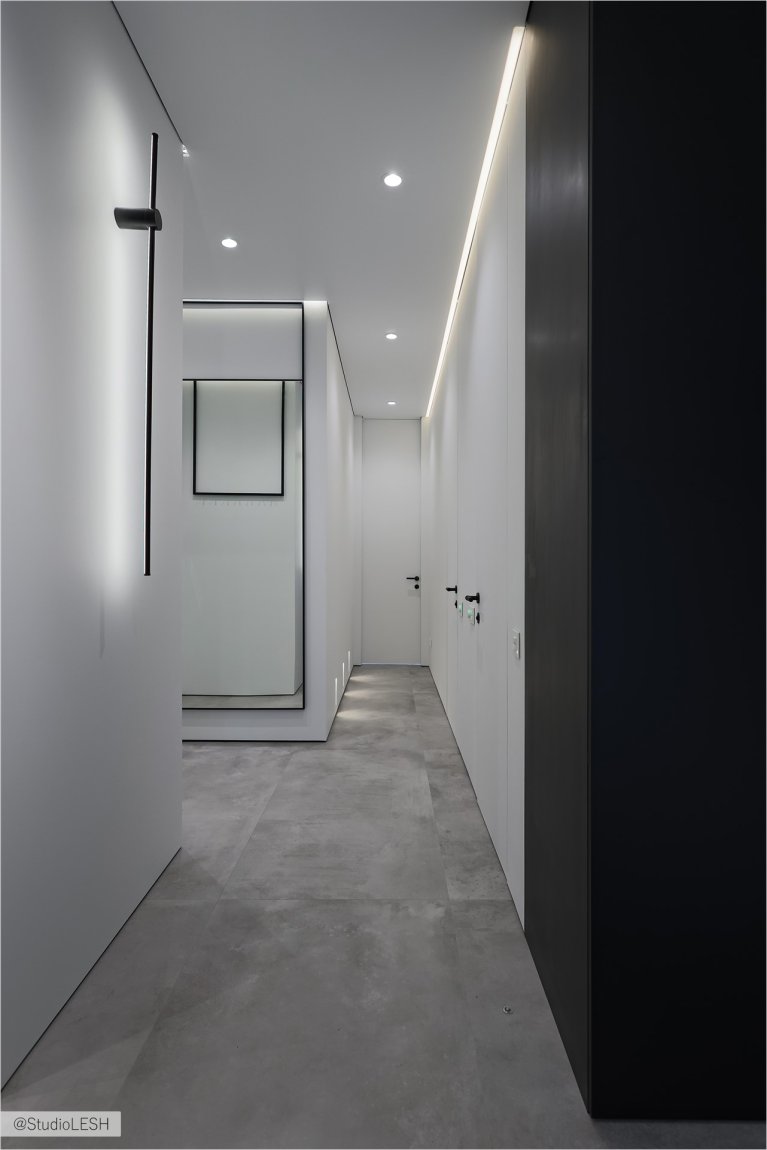 Стильный коридор с темными дверьми, подчеркнутыми светлыми стенами и потолком. На заднем плане - минималистичные светильники, создающие уютную атмосферу. Видны также другие элементы декора, такие как черная вешалка и зеркало в раме