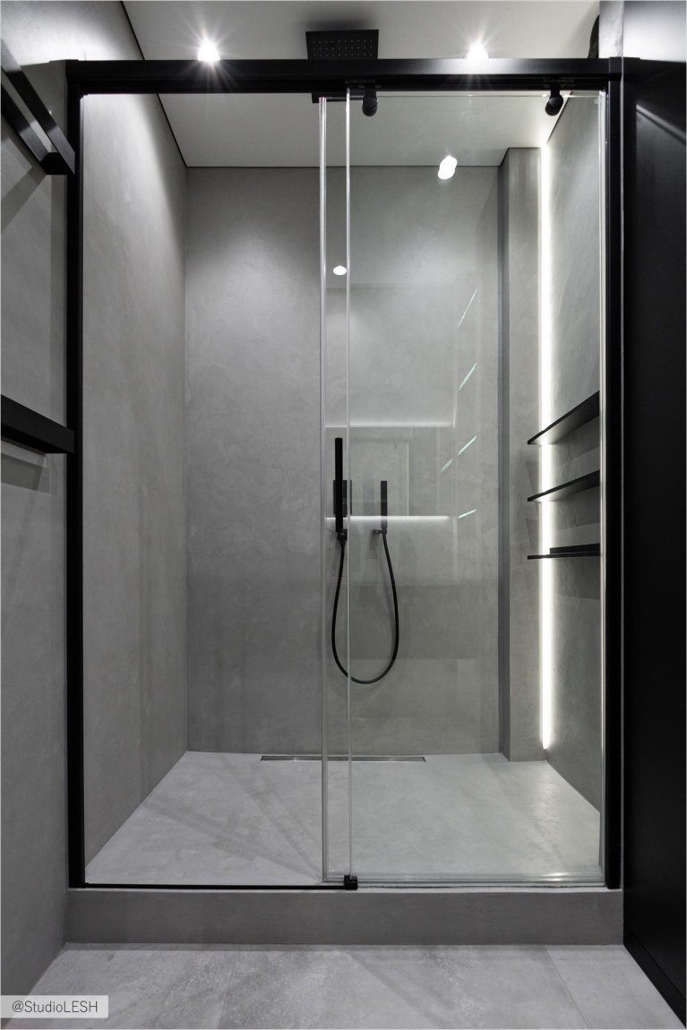 Стильная душевая кабина с большой стеклянной дверью, черным тропическим душем и минималистичными полками для банных принадлежностей. Освещение играет важную роль в создании уютной атмосферы: подсветка под потолком и на стенах подчеркивает текстуру плитки и создает уют