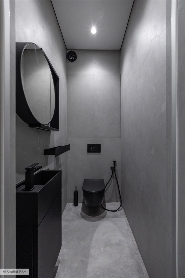 Стильный туалет с подвесным унитазом черного цвета и гигиеническим душем. Рядом с унитазом - раковина с черным смесителем и круглое зеркало в черной раме. На заднем плане - встроенные шкафы и другие элементы декора, создающие уютную атмосферу